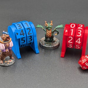 May include: Blue and red plastic counter wheels with numbers 1-6 and 0-5. A green dragon miniature and a red 20-sided die are also in the image.