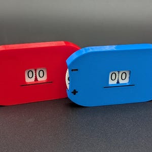 May include: Two small, rectangular counters, one red and one blue, each displaying "00" on a white background. The counters have a small wheel on the side for adjusting the numbers. The background is a dark gray.