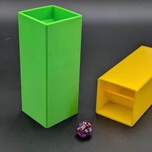 May include: A bright green, tall, rectangular container stands next to a yellow, rectangular container with an open end. A purple, multi-sided die rests between them. The objects are on a dark surface.