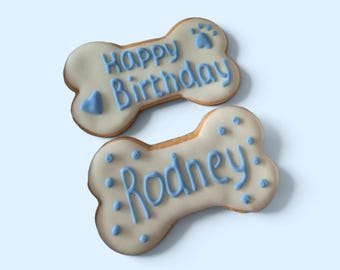 Puede incluir: Dos galletas con forma de hueso con glaseado blanco y letras en azul claro. Una galleta dice Happy Birthday con un corazón y una huella. La segunda galleta presenta el nombre Rodney rodeado de pequeños puntos azules.