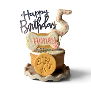 May include: A small round cake topped with colourful sprinkles, a bone-shaped biscuit with the name Honey in pink icing, a number 5 biscuit with pink dots, a black Happy Birthday cake topper, and a round biscuit embossed with Happy Birthday.