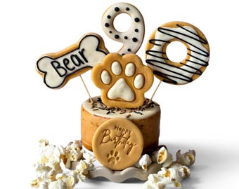 May include: A small cylindrical cake topped with a white paw print cookie, a number nine cookie, a bone-shaped cookie with the text Bear, and a donut-shaped cookie. A round cookie with the text Happy Birthday and a paw print sits in front.