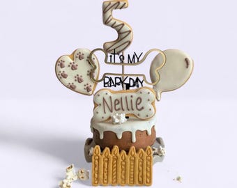 May include: A birthday cake topper set featuring a large number 5 cookie, a heart cookie with paw prints, a balloon cookie, and a bone-shaped cookie with the name Nellie. A black wire sign reads It's My Bark Day above a candle-shaped cookie base.