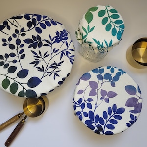 May include: Three reusable food covers with a white background and a blue and green leaf pattern. The covers are made of fabric and are designed to fit over bowls and containers.