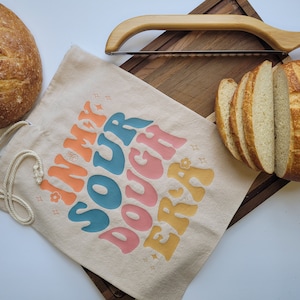 May include: A white canvas tote bag with the text "In My Sour Dough Era" printed in rainbow colors. The bag is next to a wooden cutting board with a loaf of sourdough bread and a bread knife.