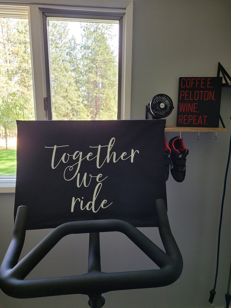 May include: Black exercise bike with a black seat cover that says "Together we ride". A small fan and a sign that says "Coffee. Peloton. Wine. Repeat." are on a shelf above the bike.