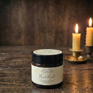 May include: A jar of Wild Hive Apothecary Muscle Mend cream with a black lid and a label. Two lit candles in brass holders are in the background. The jar is on a wooden surface.