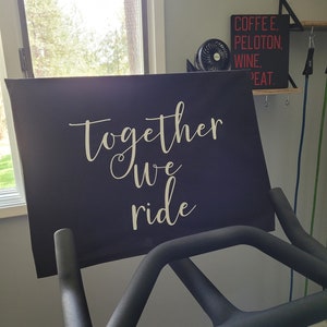 May include: A black exercise bike with a motivational sign that reads "Together we ride" in white lettering.