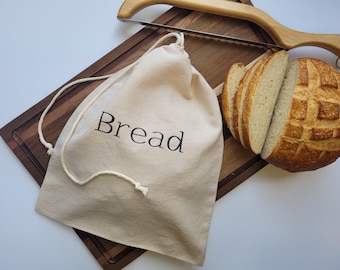 May include: A white canvas bread bag with a drawstring closure and the word "Bread" printed in black. The bag is sitting on a wooden cutting board with a loaf of bread and a wooden bread knife.