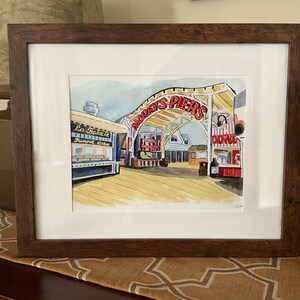 May include: A watercolor painting of a boardwalk scene with a red and white sign that reads "Doddys Piers". The painting features a blue and white building with the text "La Bakerie, Funnel Cakes" and other shops and buildings in the background.
