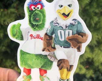 May include: A die-cut sticker featuring a watercolor illustration of two sports mascots standing side-by-side. The left mascot is a fuzzy green creature in a white jersey with the word Phillie in red script. The right mascot is a white eagle.