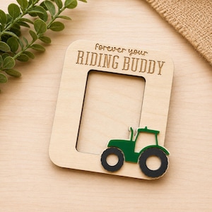 May include: A rectangular light-colored wooden picture frame with rounded corners. The top features the engraved text "forever your RIDING BUDDY". A green and black tractor cutout is attached to the bottom right corner of the frame.