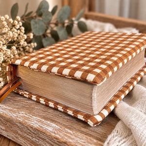 May include: A rectangular book cover made of fabric with a brown and white gingham check pattern. The cover features a padded texture and includes two thin brown ribbon bookmarks extending from the side of the closed book.