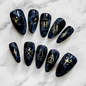 Puede incluir: Conjunto de diez uñas postizas negras y doradas con un diseño de mármol y varios símbolos temáticos de Harry Potter, incluyendo las Reliquias de la Muerte, el escudo de Hogwarts, la Snitch Dorada y el letrero de la Plataforma 9 3/4.