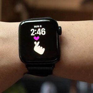 May include: A black smartwatch displaying the time 2:46, the day of the week, and a heart symbol with a hand gesture. The watch is worn on a wrist with a black band.