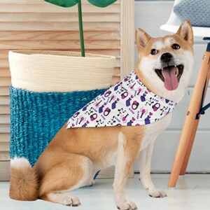 May include: A Shiba Inu dog wearing a white bandana printed with purple and pink K-Pop themed graphics. The dog is sitting with its tongue out. A blue woven basket and a wooden stool are in the background.