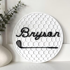 May include: A circular white wall sign featuring a textured honeycomb pattern resembling a golf ball. The sign displays the name Bryson in black cursive script lettering, positioned above a black silhouette of a golf club iron.
