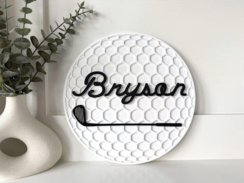 May include: A circular white wall sign featuring a textured honeycomb pattern resembling a golf ball. The sign displays the name Bryson in black cursive script lettering, positioned above a black silhouette of a golf club iron.