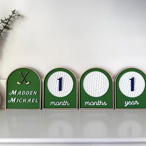 May include: Four arched wooden signs with green backgrounds. The first features two golf clubs and the text Madden Michael. The others display white golf balls with the numbers 1 and the text month, months, and year in white script lettering.