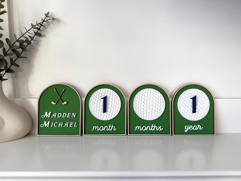 May include: Four arched wooden signs with green backgrounds. The first features two golf clubs and the text Madden Michael. The others display white golf balls with the numbers 1 and the text month, months, and year in white script lettering.