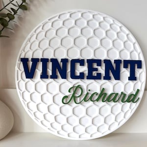 May include: White golf ball-shaped wall decor with a textured surface and a honeycomb pattern. The name "VINCENT" is displayed in navy blue letters, and "Richard" is in green script. Decorative item for a golf enthusiast.