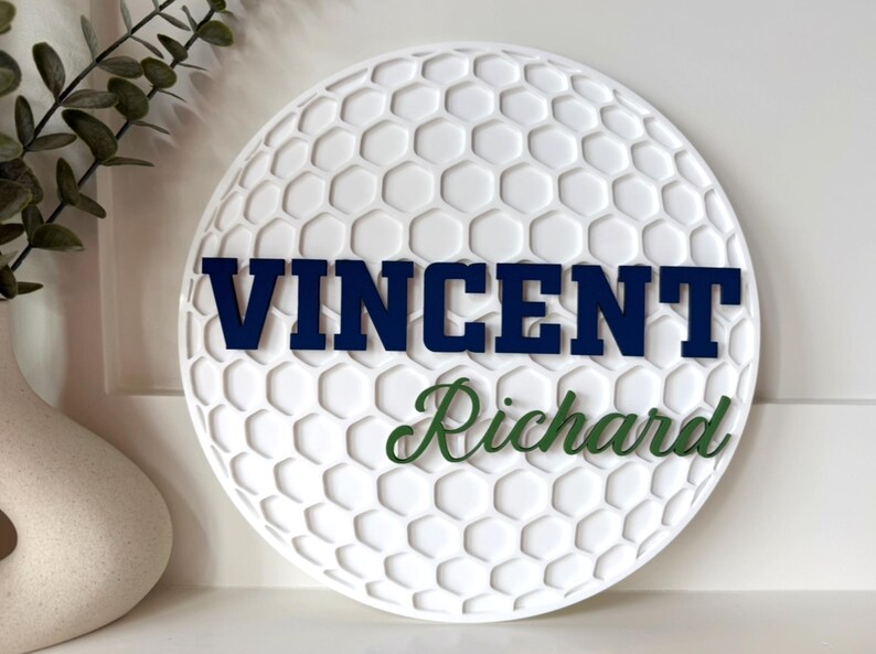 May include: White golf ball-shaped wall decor with a textured surface and a honeycomb pattern. The name "VINCENT" is displayed in navy blue letters, and "Richard" is in green script. Decorative item for a golf enthusiast.