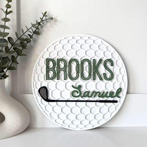 May include: A circular white wall sign with a textured golf ball dimple pattern. The sign features the name BROOKS in dark green block letters, the name Samuel in dark green script, and a black golf club graphic positioned horizontally.
