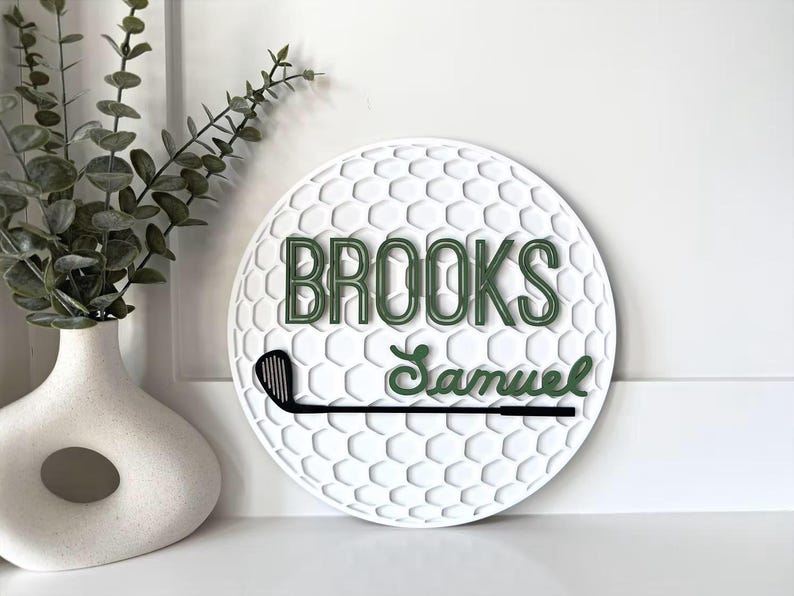 May include: A circular white wall sign with a textured golf ball dimple pattern. The sign features the name BROOKS in dark green block letters, the name Samuel in dark green script, and a black golf club graphic positioned horizontally.