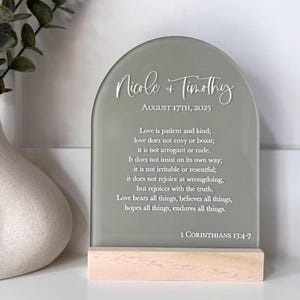 May include: A sage green arched glass wedding sign with the names "Nicole & Timothy" and the date "August 17th, 2025" in white script. The sign includes a Bible verse and sits on a light wood base.