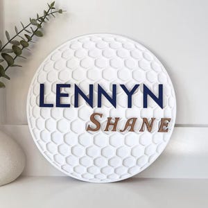 May include: A white, round decorative sign with a golf ball texture. The name "LENNYN" is in navy blue letters above the name "SHANE" in brown letters. A white vase with greenery is to the left.