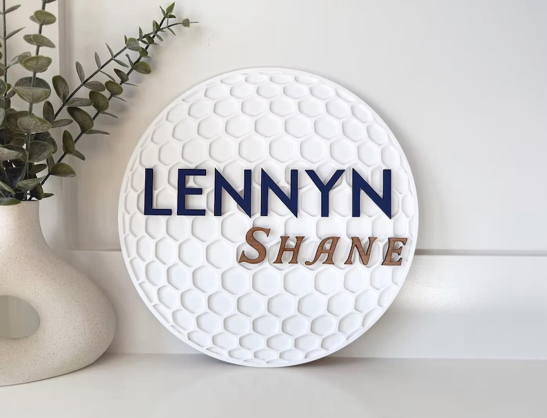 May include: A white, round decorative sign with a golf ball texture. The name "LENNYN" is in navy blue letters above the name "SHANE" in brown letters. A white vase with greenery is to the left.