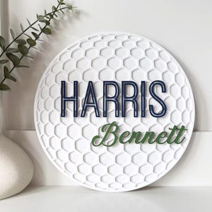 May include: A white, round decorative sign with a golf ball pattern. The sign features the name "Harris" in navy blue and "Bennett" in green. A vase with greenery is in the background.