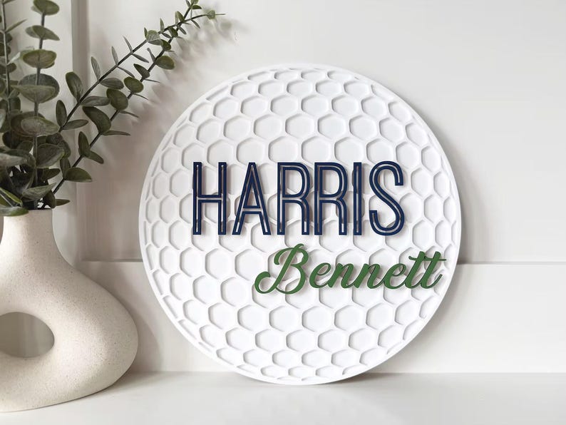 May include: A white, round decorative sign with a golf ball pattern. The sign features the name "Harris" in navy blue and "Bennett" in green. A vase with greenery is in the background.