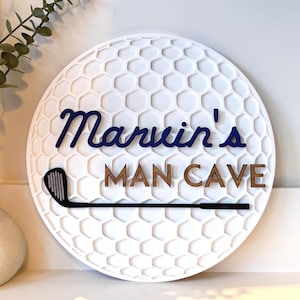 May include: A circular white wall sign with a textured honeycomb pattern resembling a golf ball. The sign features the dark blue script text Marvin's and the brown block text MAN CAVE, with a black golf club graphic positioned horizontally below.