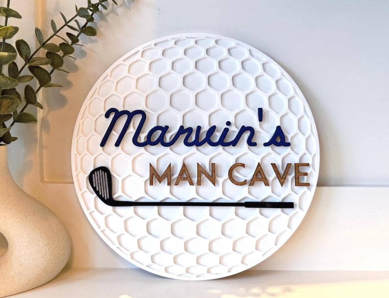 May include: A circular white wall sign with a textured honeycomb pattern resembling a golf ball. The sign features the dark blue script text Marvin's and the brown block text MAN CAVE, with a black golf club graphic positioned horizontally below.