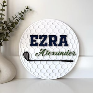May include: A circular white wall sign with a textured golf ball dimple pattern. The sign features the name EZRA in bold navy blue block letters, the name Alexander in green script, and a black silhouette of a golf club across the center.