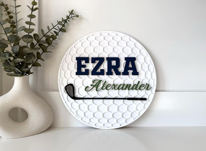 May include: A circular white wall sign with a textured golf ball dimple pattern. The sign features the name EZRA in bold navy blue block letters, the name Alexander in green script, and a black silhouette of a golf club across the center.