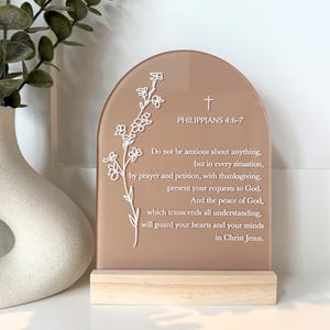 May include: A decorative sign with a light brown arched top and a wooden base. The sign features a white floral design and text from Philippians 4:6-7. The text reads, "Do not be anxious about anything..."