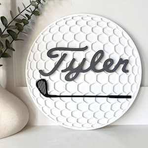May include: A circular white wall sign featuring a textured honeycomb pattern resembling a golf ball. The name Tyler is written in dark gray cursive script across the center, positioned above a black silhouette of a golf club iron.