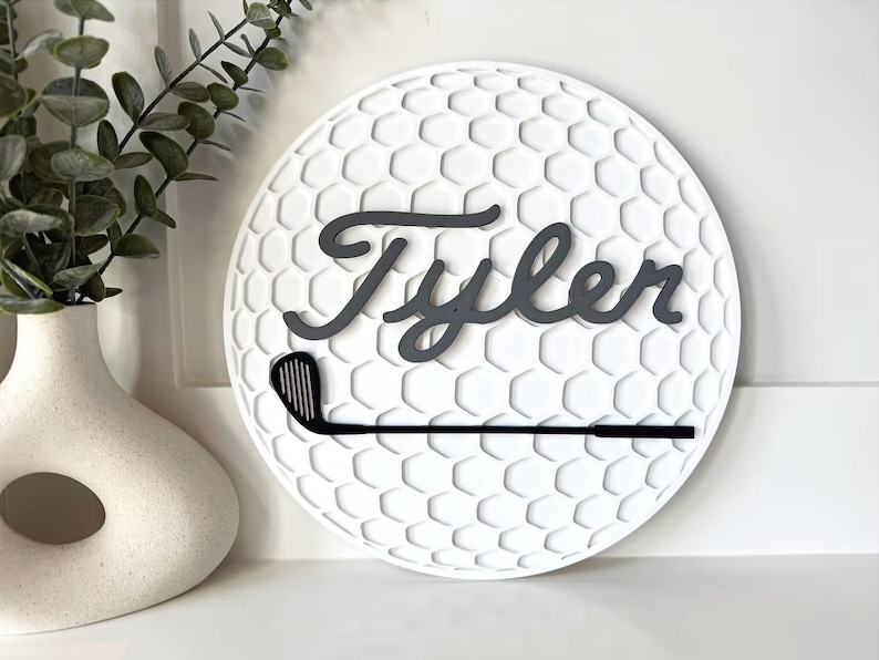 May include: A circular white wall sign featuring a textured honeycomb pattern resembling a golf ball. The name Tyler is written in dark gray cursive script across the center, positioned above a black silhouette of a golf club iron.