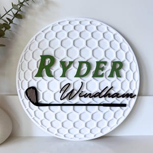 May include: A circular white wall sign featuring a textured honeycomb pattern resembling a golf ball. The sign displays the name RYDER in green block letters and Windham in black script, with a black golf club graphic positioned below.