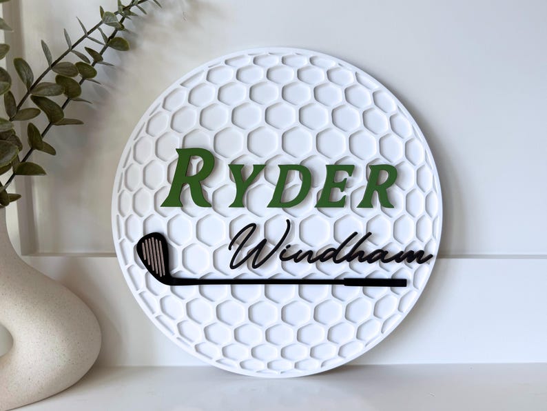 May include: A circular white wall sign featuring a textured honeycomb pattern resembling a golf ball. The sign displays the name RYDER in green block letters and Windham in black script, with a black golf club graphic positioned below.