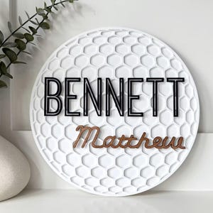 May include: A white, round decorative sign with a golf ball texture. The name "BENNETT" is in black, outlined letters above the name "Matthew" in a brown, cursive font. A white vase with greenery is to the left.