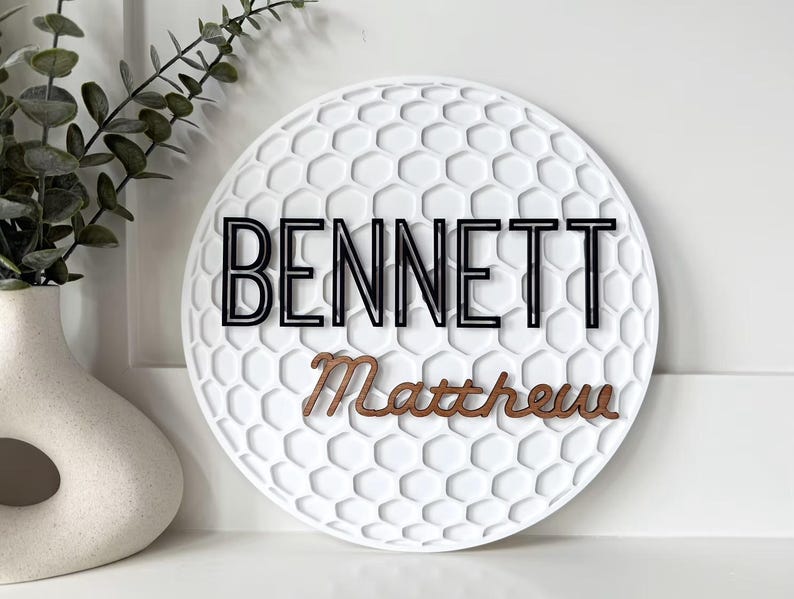 May include: A white, round decorative sign with a golf ball texture. The name "BENNETT" is in black, outlined letters above the name "Matthew" in a brown, cursive font. A white vase with greenery is to the left.