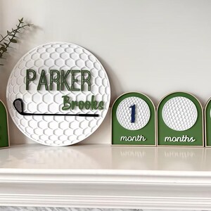 May include: A circular white golf ball textured sign with the name Parker Brooks and a black golf club, alongside a green arched sign with crossed golf clubs and three milestone signs featuring golf balls and the text 1 month, months, and 1 year.