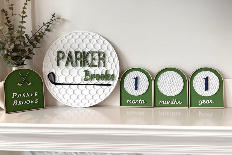 May include: A circular white golf ball textured sign with the name Parker Brooks and a black golf club, alongside a green arched sign with crossed golf clubs and three milestone signs featuring golf balls and the text 1 month, months, and 1 year.