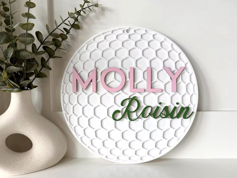 May include: White round wall decor with a honeycomb pattern and the name "MOLLY" in pink letters and "Roisin" in green script. A decorative vase with greenery is to the left.