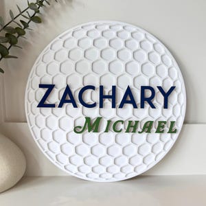 May include: A white, round sign with a golf ball pattern. The name "ZACHARY" is in navy blue letters above "MICHAEL" in green. The sign is a decorative item.