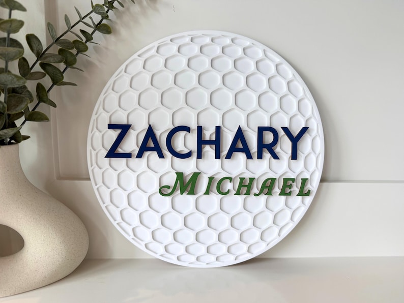 May include: A white, round sign with a golf ball pattern. The name "ZACHARY" is in navy blue letters above "MICHAEL" in green. The sign is a decorative item.