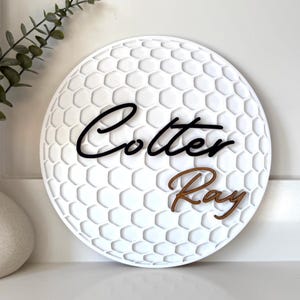 May include: White, round wall decor with a golf ball pattern. The name "Cotter Ray" is displayed in black and brown script. Decorative item for a home.